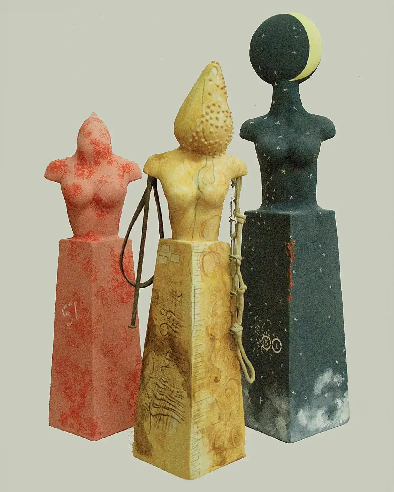 Three tall totemic ceramic figures side by side in coral, ochre and deep teal, each with a stylised torso and an elongated crowned head.
